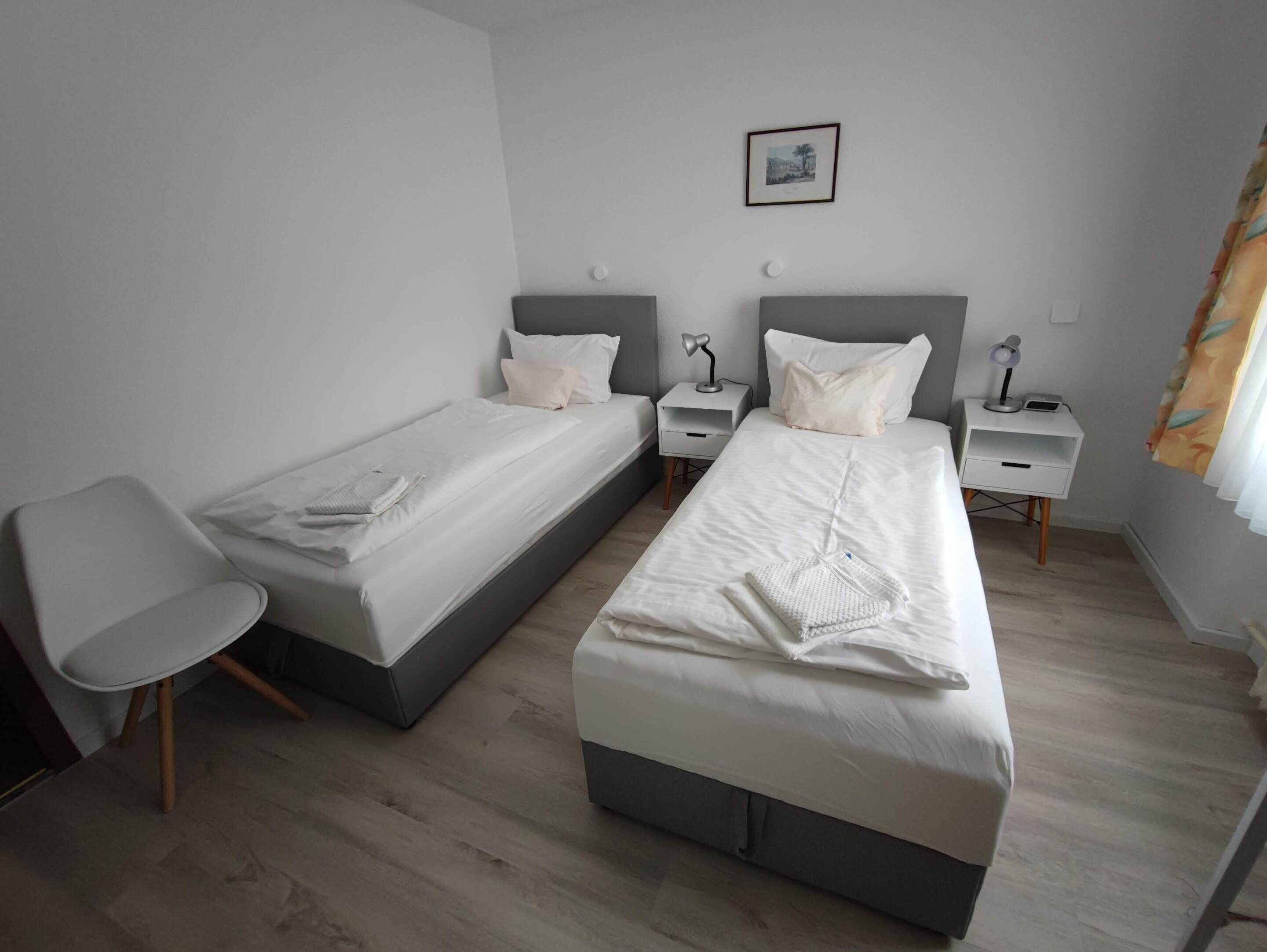 Et moderne hotelværelse med to senge, et lille skrivebord, en stol og et natbord med lampe.