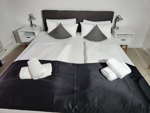 Una cama doble impecablemente arreglada con ropa de cama blanca y gris, dos mesitas de noche con lámparas y un corredor de cama negro decorativo.