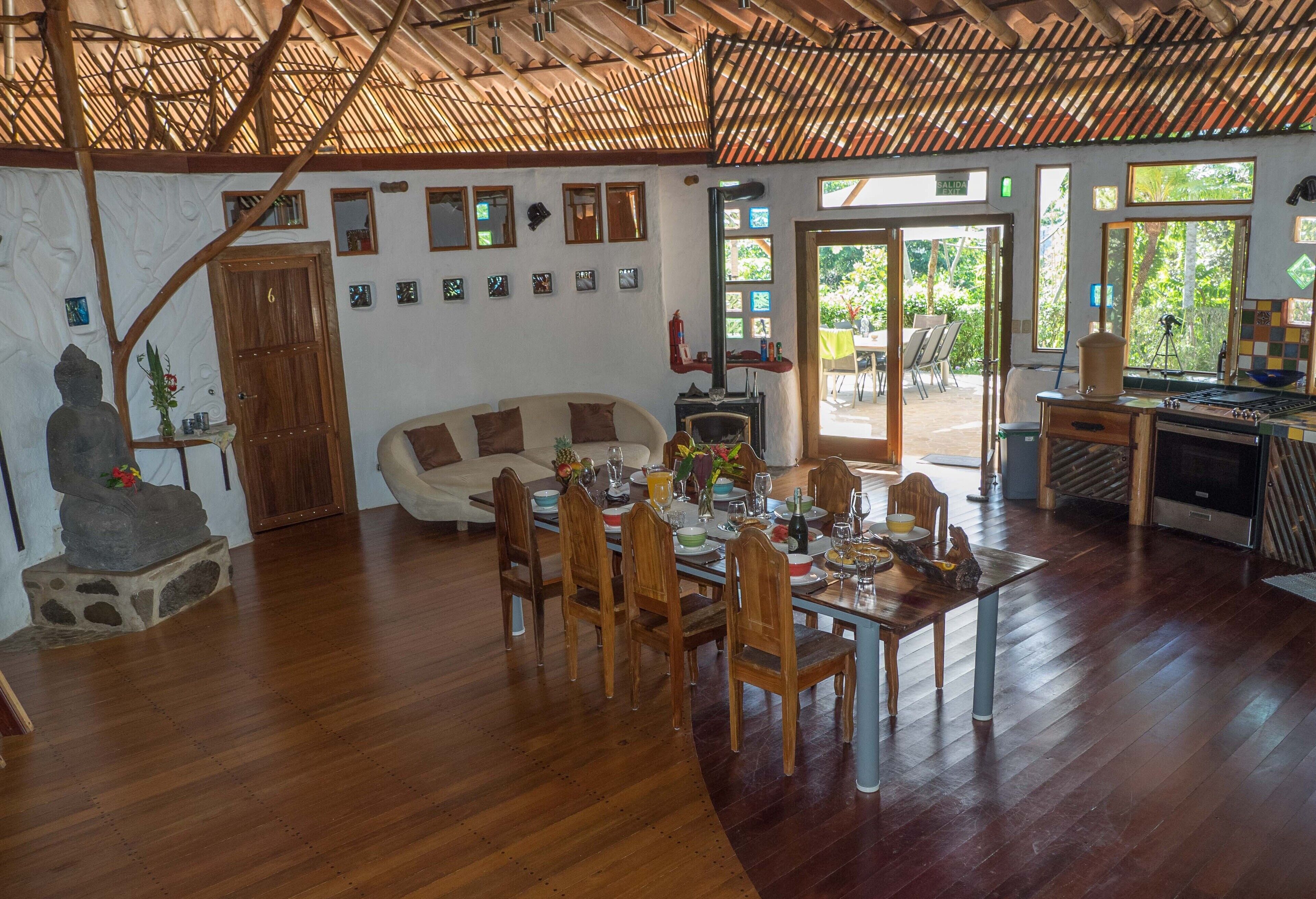 A spacious living area with a wooden dining table set for six, surrounded by wooden chairs. A sofa and a stone Buddha statue are also visible.