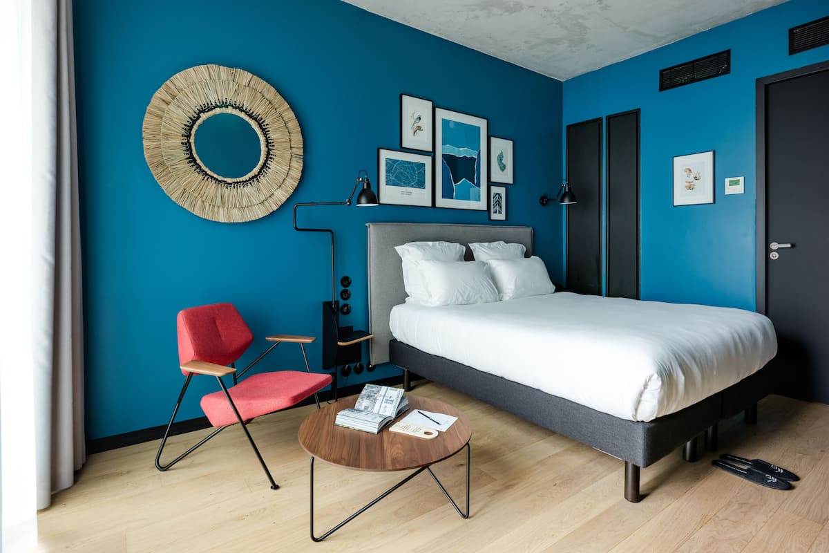 Un dormitorio moderno con una cama grande, una silla roja, una mesa de centro redonda de madera y obras de arte en las paredes.