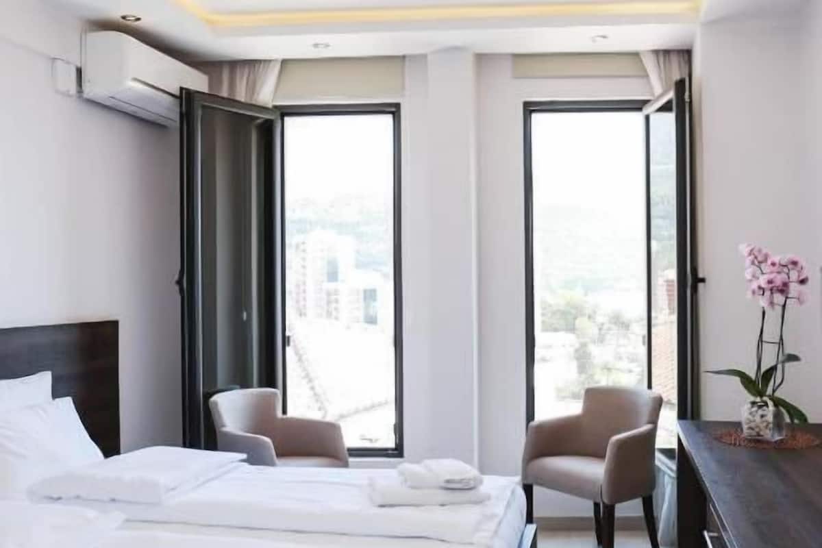 Ein modernes Hotelzimmer mit einem großen Bett, zwei Sesseln, einer Kommode und Blick auf die Stadt durch große Fenster.