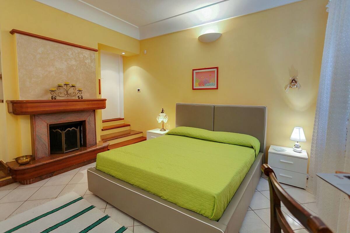A bedroom with a bed, a nightstand, a lamp, a fireplace, and a picture on the wall.