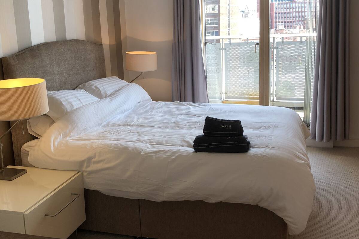 Ein Hotelzimmer mit einem großen Bett, einem Nachttisch mit Lampe, einer Kopfteil und Blick auf die Stadt durch das Fenster.