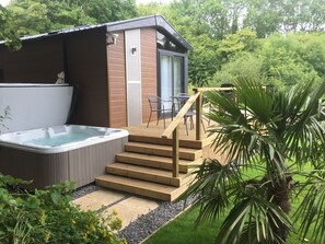 Un bain à remous situé à côté d’une terrasse en bois, accessible par des marches menant à une porte vitrée, le tout entouré de verdure.