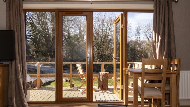 A room with a wooden floor, a dining table with chairs, a TV, and a sliding glass door leading to an outdoor deck with a wooden chair and a tree stump. - Herons Lake Retreat (Mold)