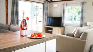 A living room with a wooden table, a bottle of champagne, a plate of strawberries, a sofa, and a television. - Herons Lake Retreat (Mold)