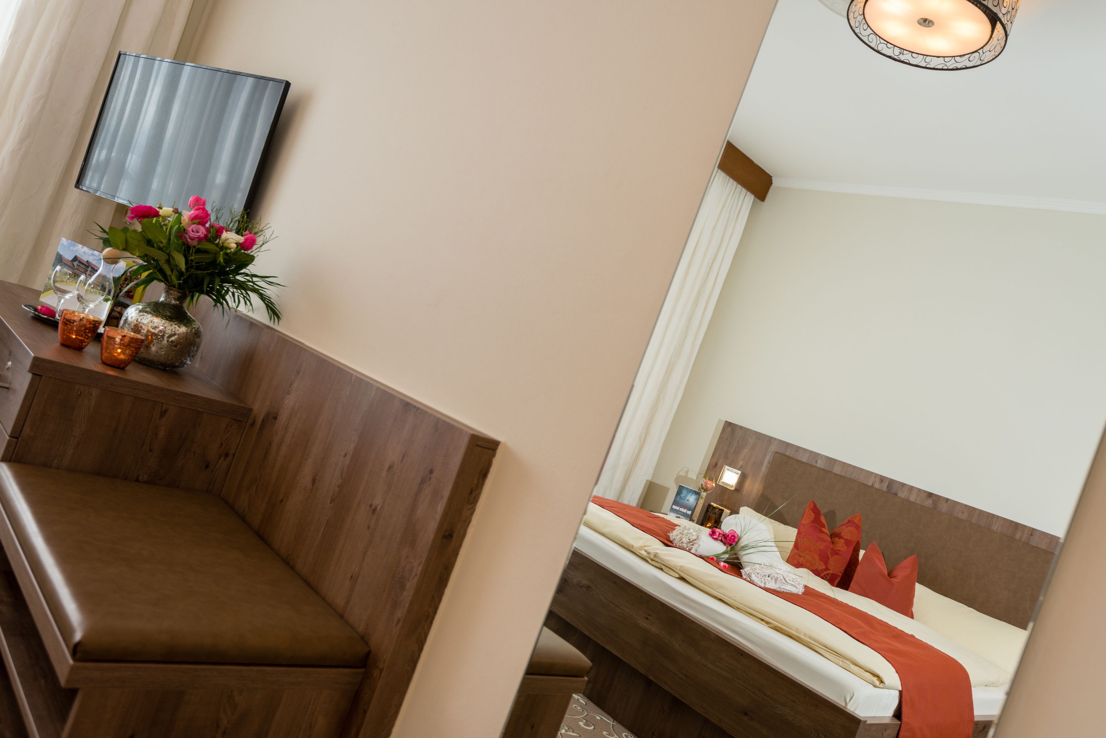 Hotelkamer met een houten bankje, nachtkastje met een bloevas, bed met rode kussens en een spiegel die de kamer weerspiegelt.