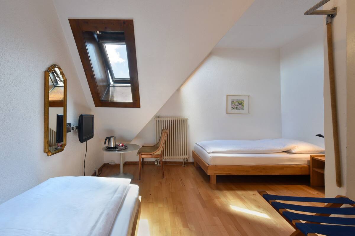 Une petite pièce bien éclairée, avec un plafond en pente, un lit simple, une chaise, une petite table avec une bouilloire et un radiateur.