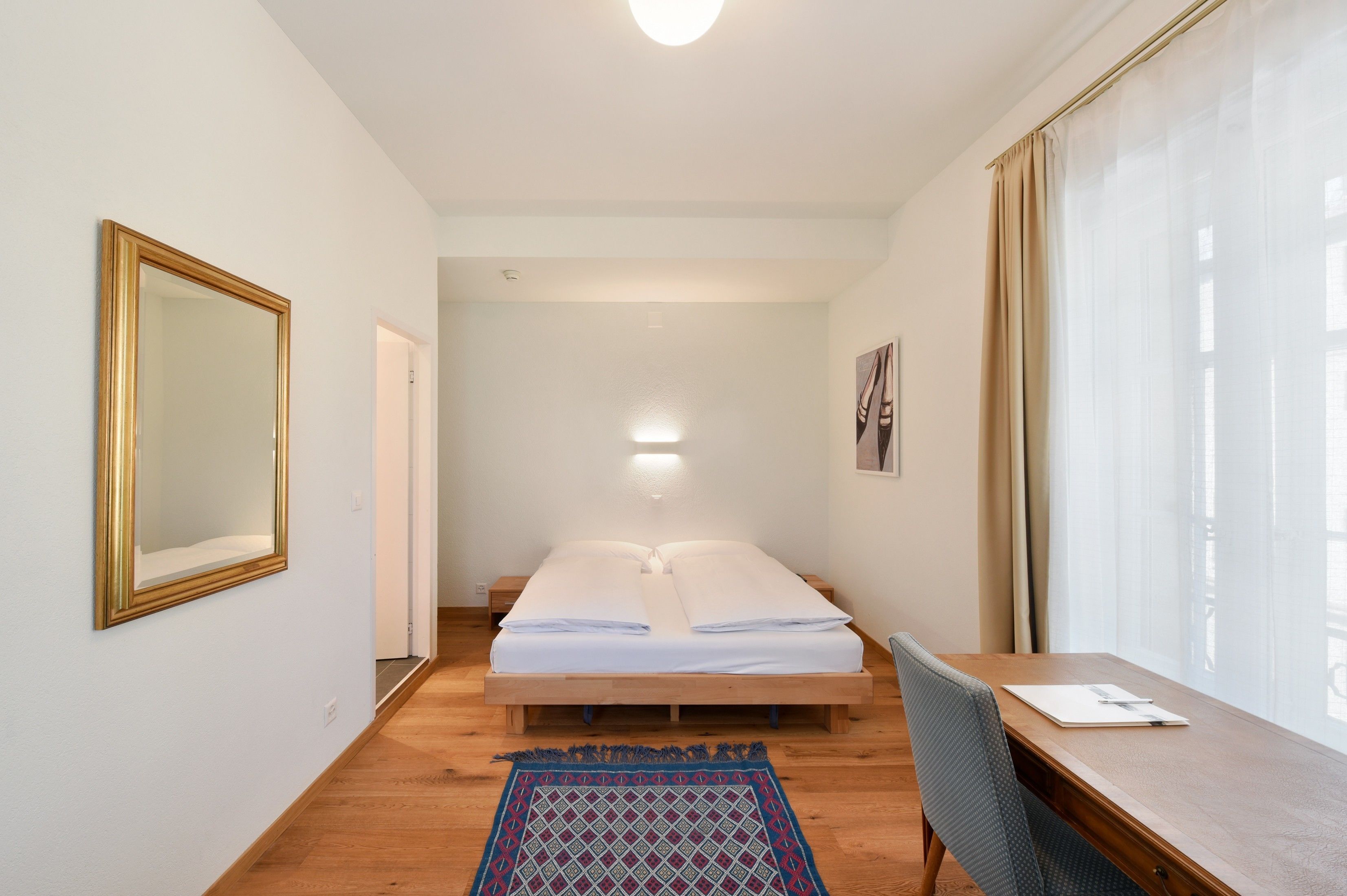 Ein Schlafzimmer mit einem Bett, einem Schreibtisch, einem Stuhl, einem Spiegel und einem Fenster mit Vorhängen.