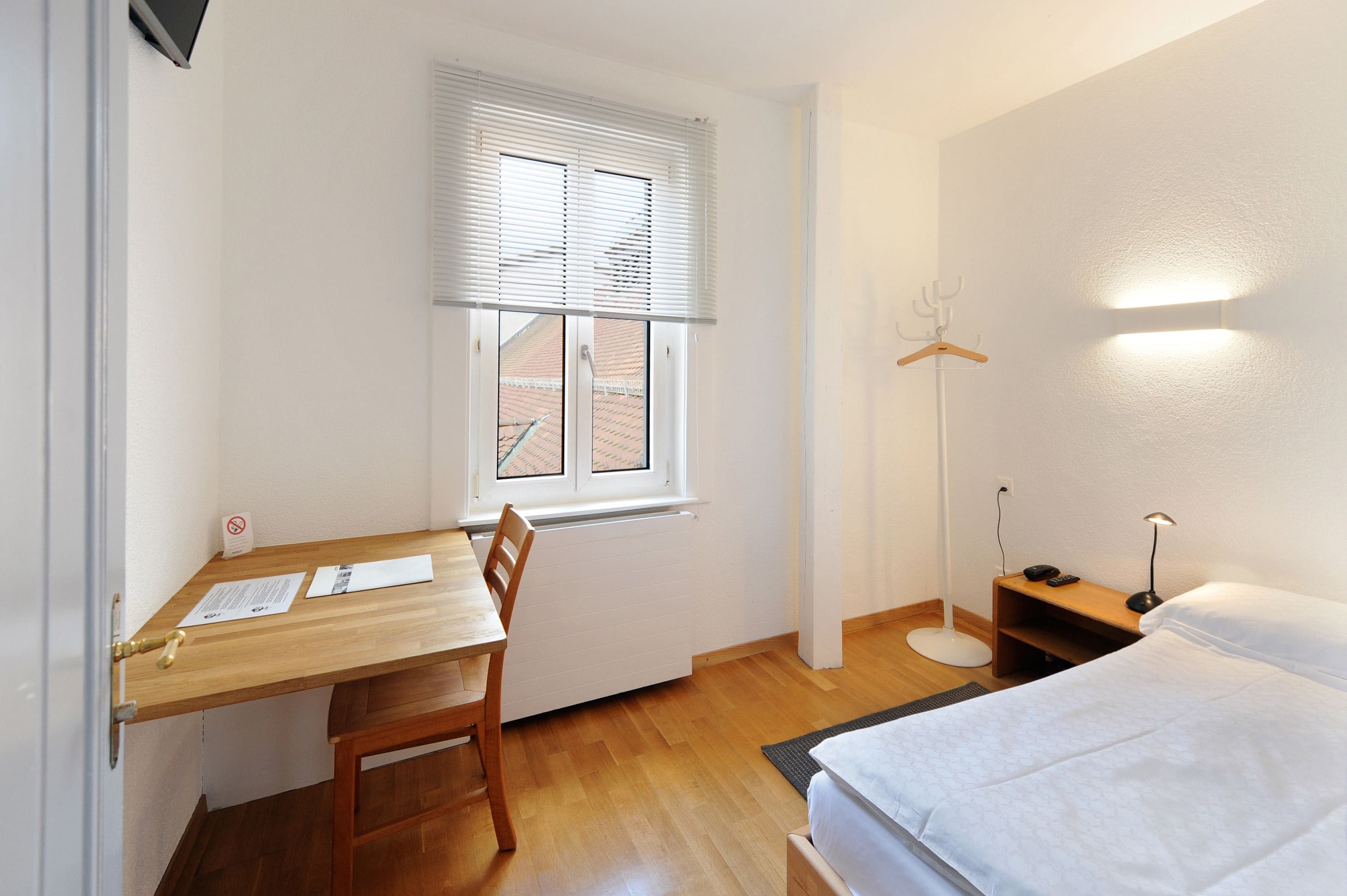 A small, clean room with a wooden desk, a bed, a floor lamp, and a window with blinds.