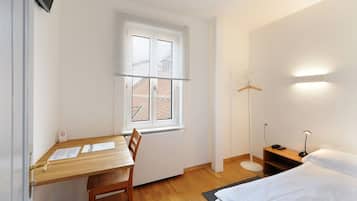 A small, clean room with a wooden desk, a bed, a floor lamp, and a window with blinds.
