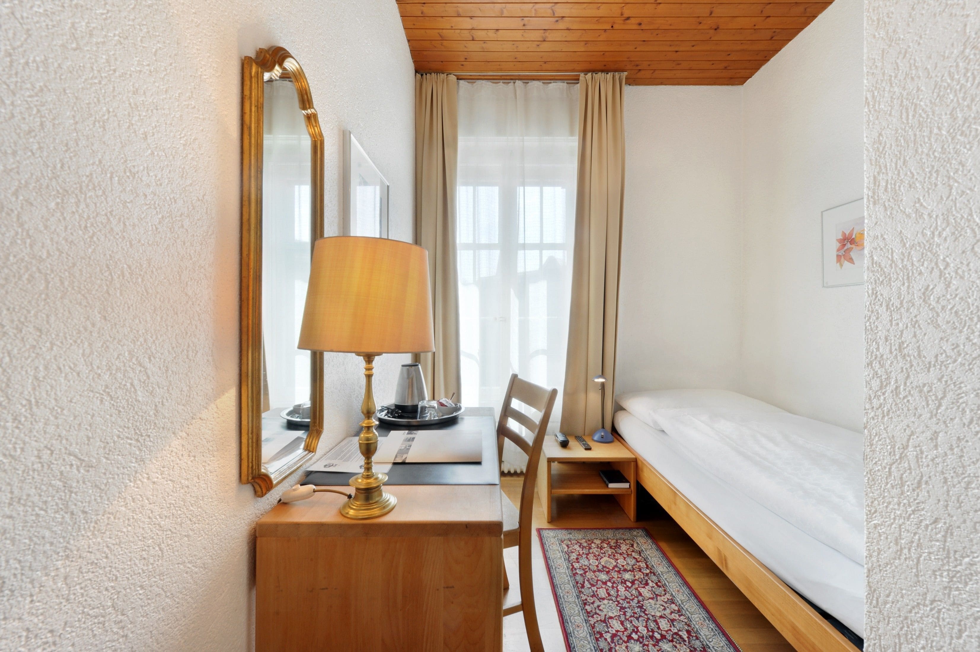 A small, cozy room with a wooden desk, a lamp, a bed, and a window with curtains.