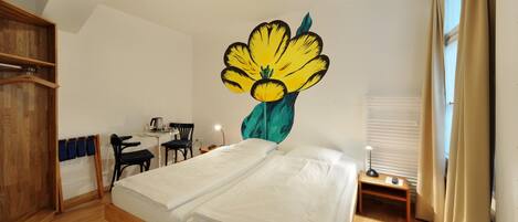 A bedroom with two single beds, a wooden wardrobe, a desk with a chair, a small table with a lamp, and a large wall mural of a flower.