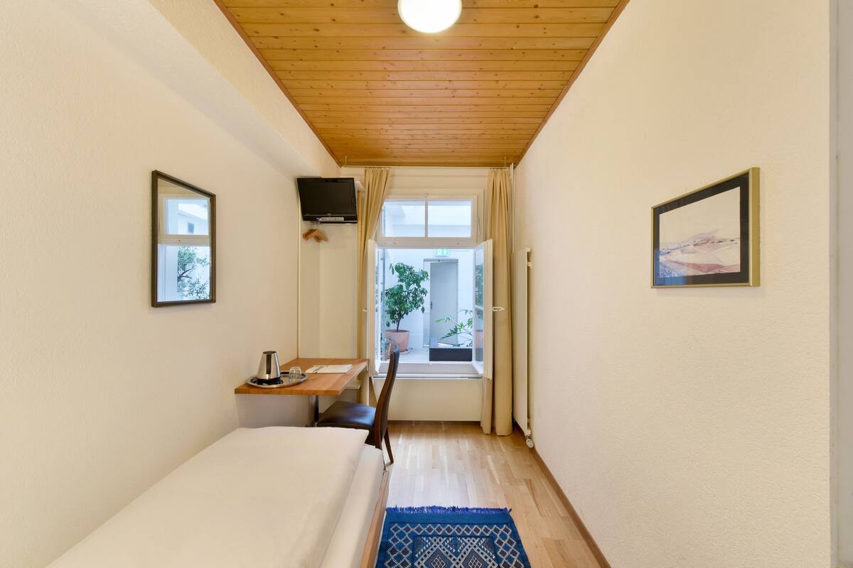 Une chambre d’hôtel étroite avec un plafond en bois, un lit, un bureau avec une chaise, un téléviseur fixé au mur et une fenêtre avec des rideaux.