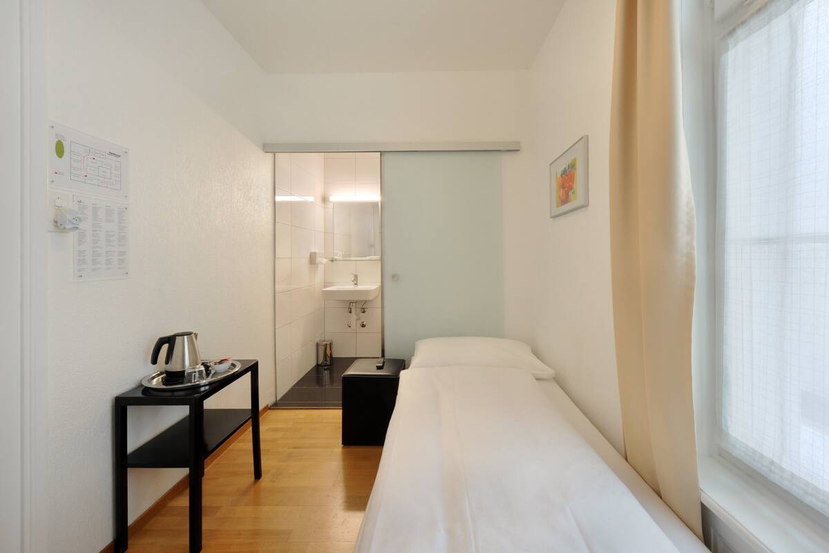 Une petite pièce propre, avec un lit, une table sur laquelle se trouve une bouilloire, un lavabo et une fenêtre avec des rideaux.