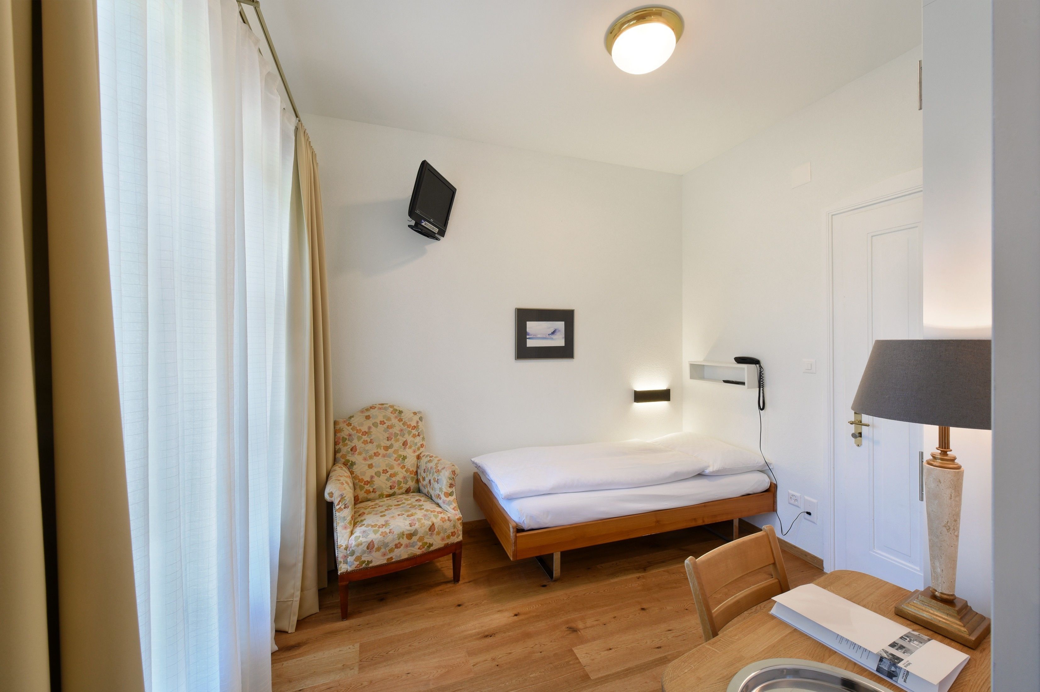 Una stanza piccola e pulita con un letto singolo, una sedia, una scrivania con una lampada e una televisione a muro.