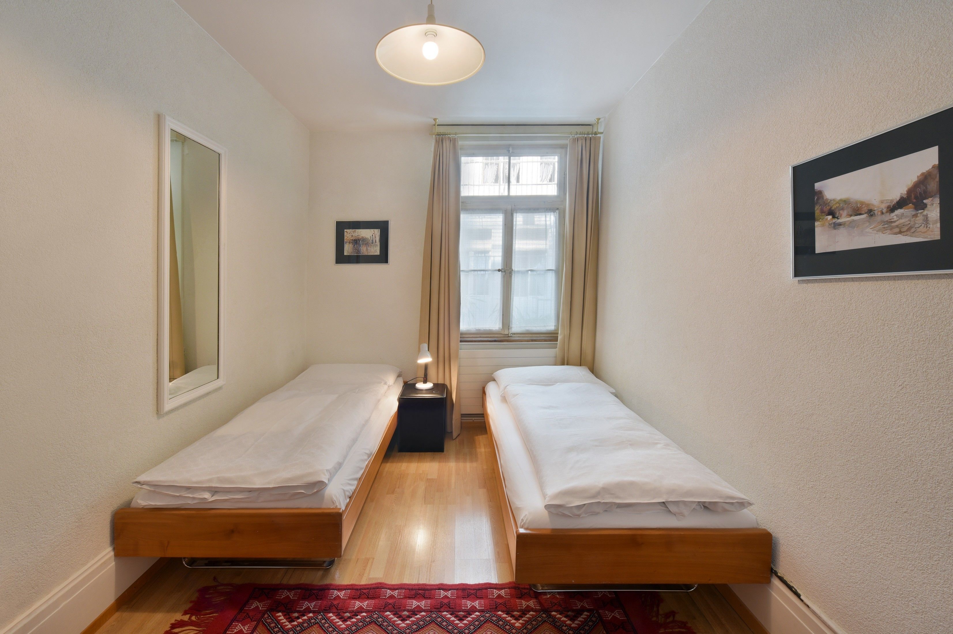 Una habitación pequeña y limpia con dos camas individuales, una mesita, una lámpara y una ventana con cortinas.
