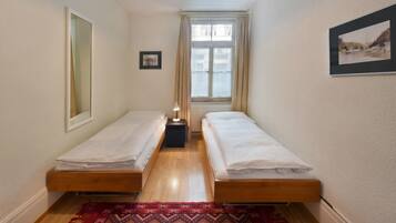 Una habitación pequeña y limpia con dos camas individuales, una mesita, una lámpara y una ventana con cortinas.