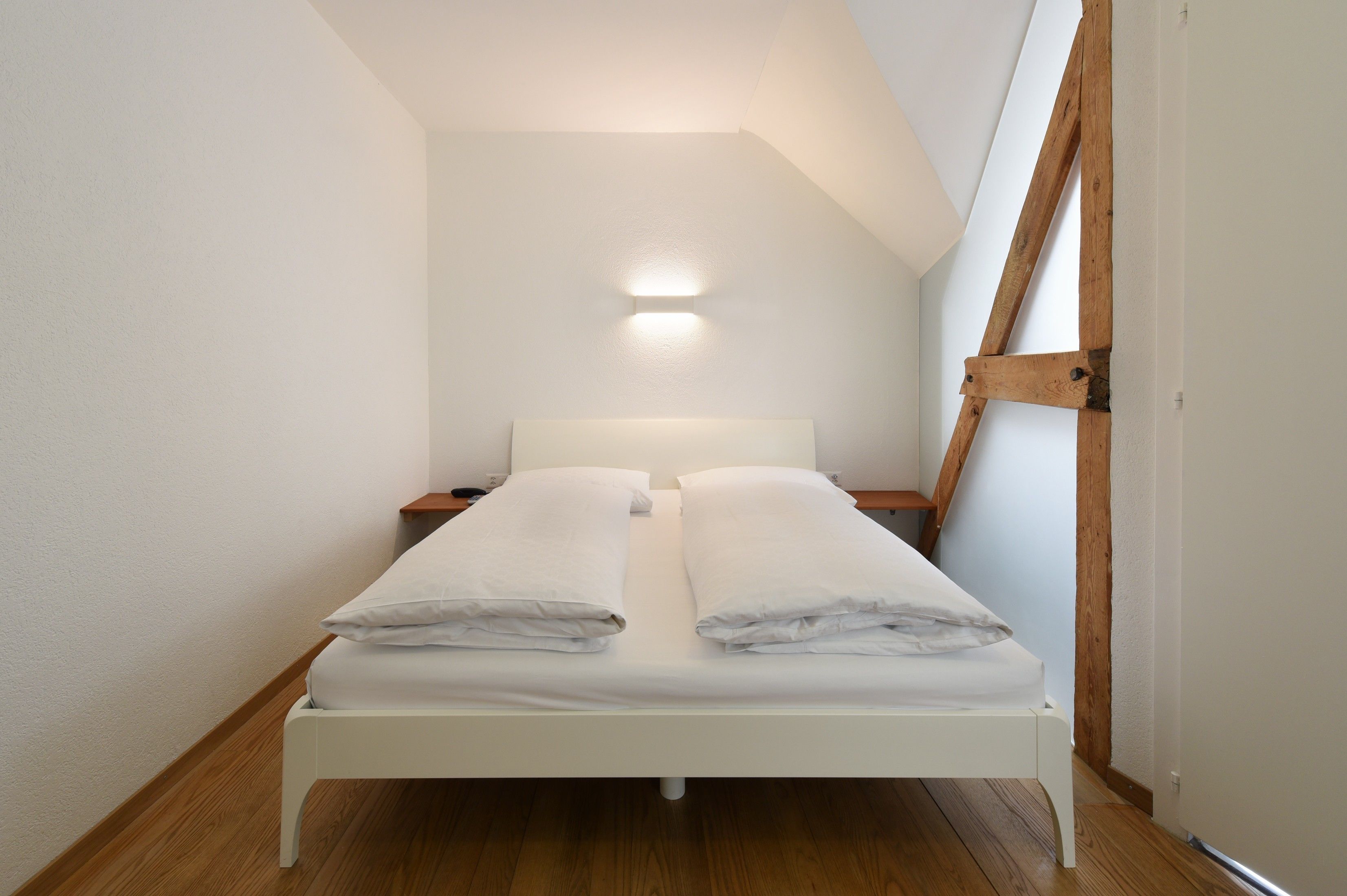 A small, white, single-bed room with a wooden floor, a nightstand, and a wall-mounted light.