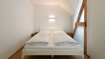 A small, white, single-bed room with a wooden floor, a nightstand, and a wall-mounted light.