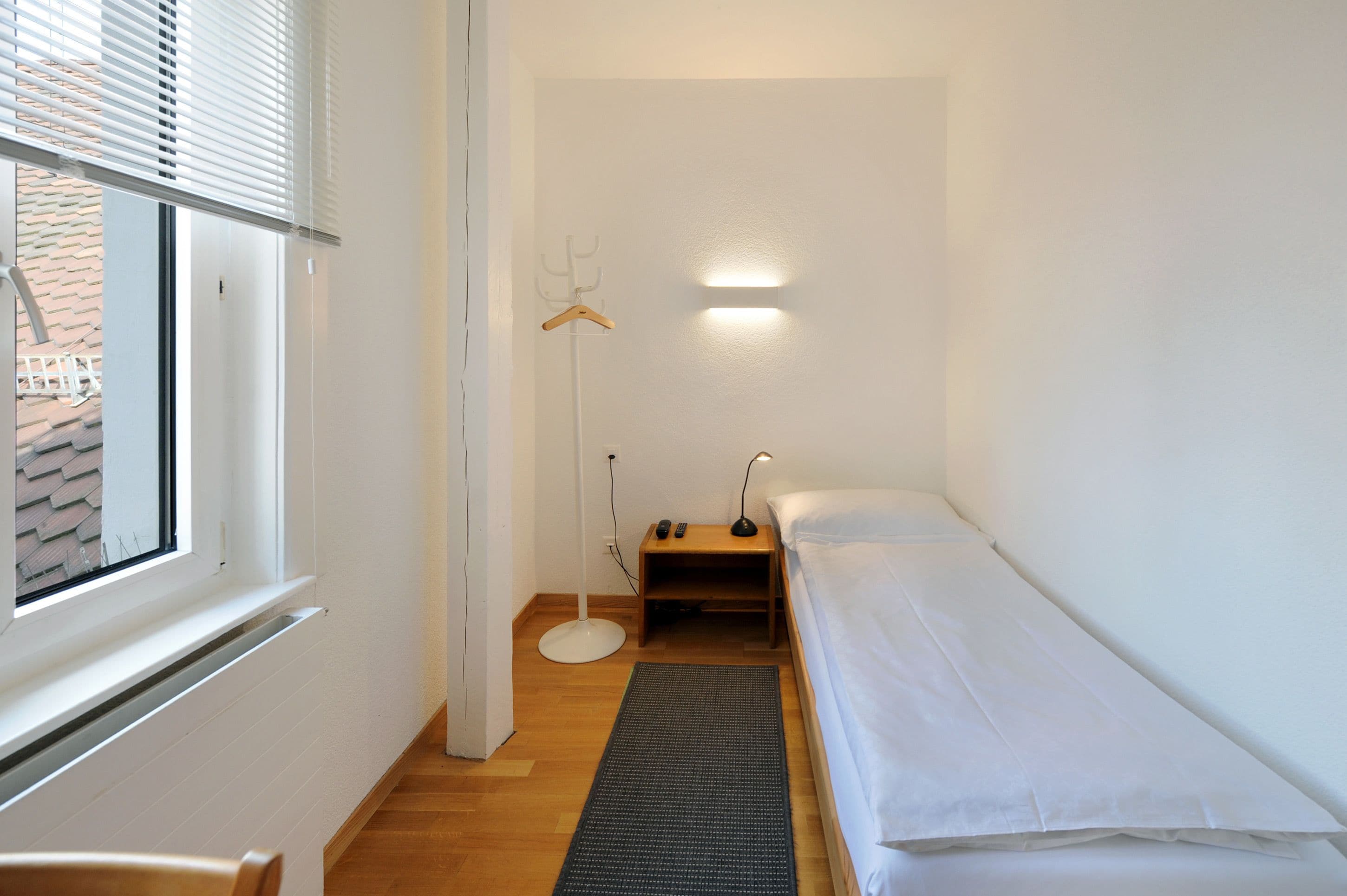 Una stanza piccola e pulita con un letto singolo, una lampada da terra e un attaccapanni.