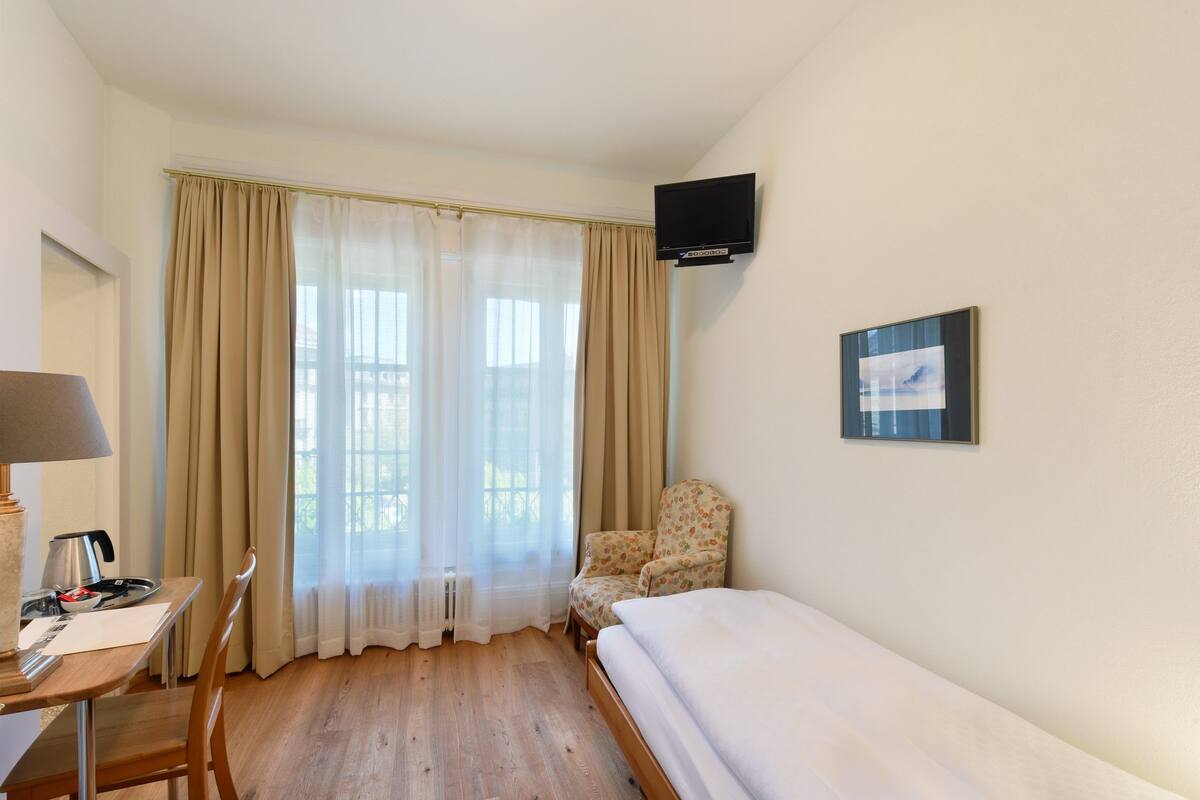 Habitación de hotel con cama, una silla, escritorio con lámpara, televisor y ventana con cortinas.