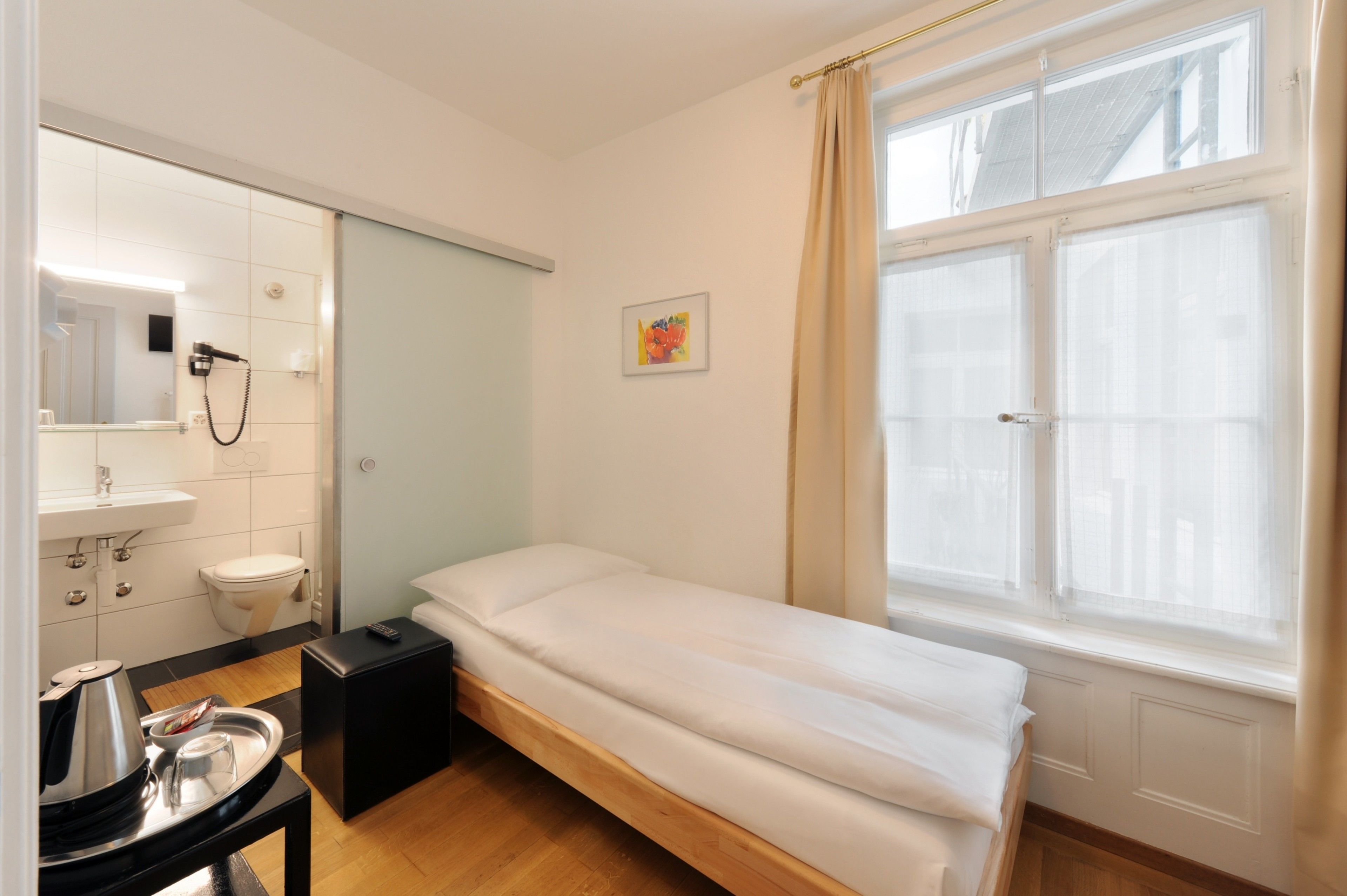 Una habitación pequeña y limpia con una cama individual, una mesita de noche con lámpara, una ventana con cortinas y un baño con espejo y lavabo.