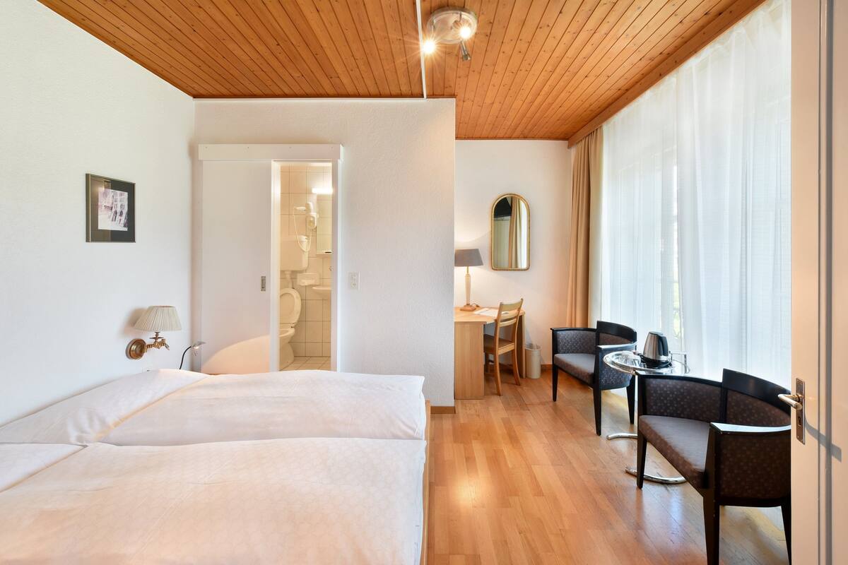 Une chambre d’hôtel avec deux lits, un bureau, une chaise, une lampe et une grande fenêtre avec des rideaux.
