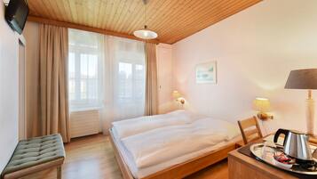A hotel room with a wooden ceiling, a bed with white linens, a bench, a desk with a lamp, and a window with curtains.