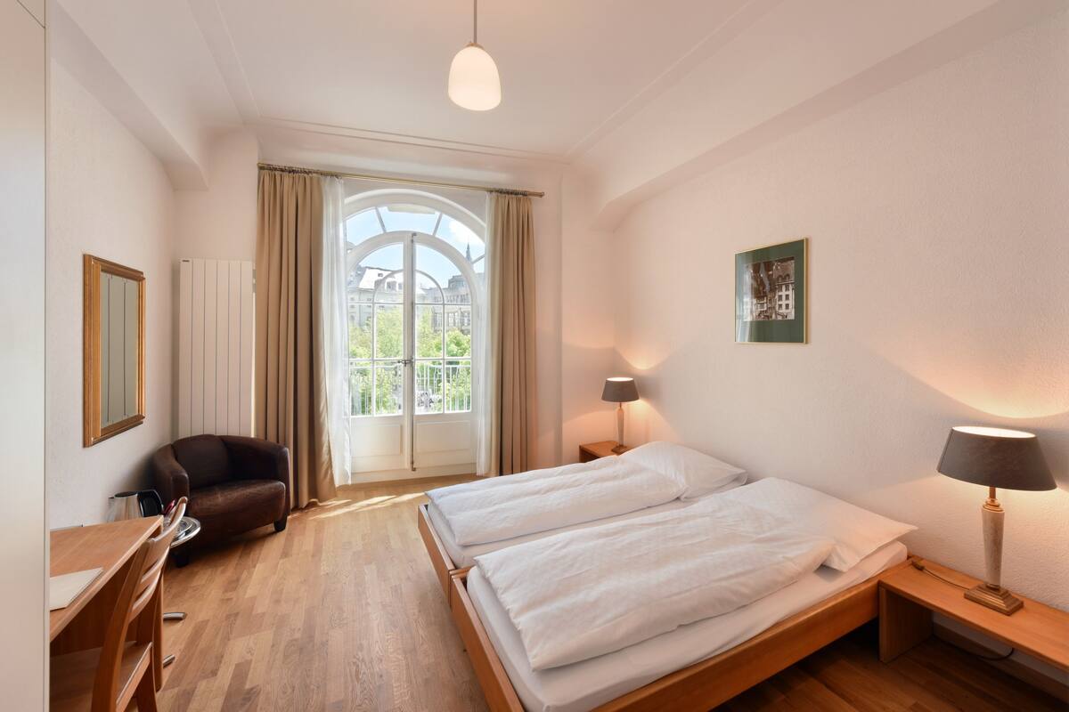 Une chambre d’hôtel avec un lit, un bureau, une chaise, une lampe et une fenêtre avec des rideaux.