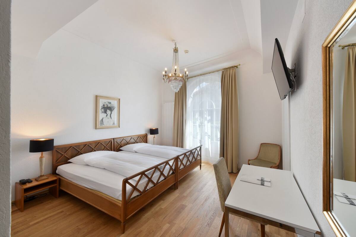 Une chambre d’hôtel avec un lit en bois, un bureau, une chaise, un lustre, un téléviseur à écran plat et un tableau encadré au mur.