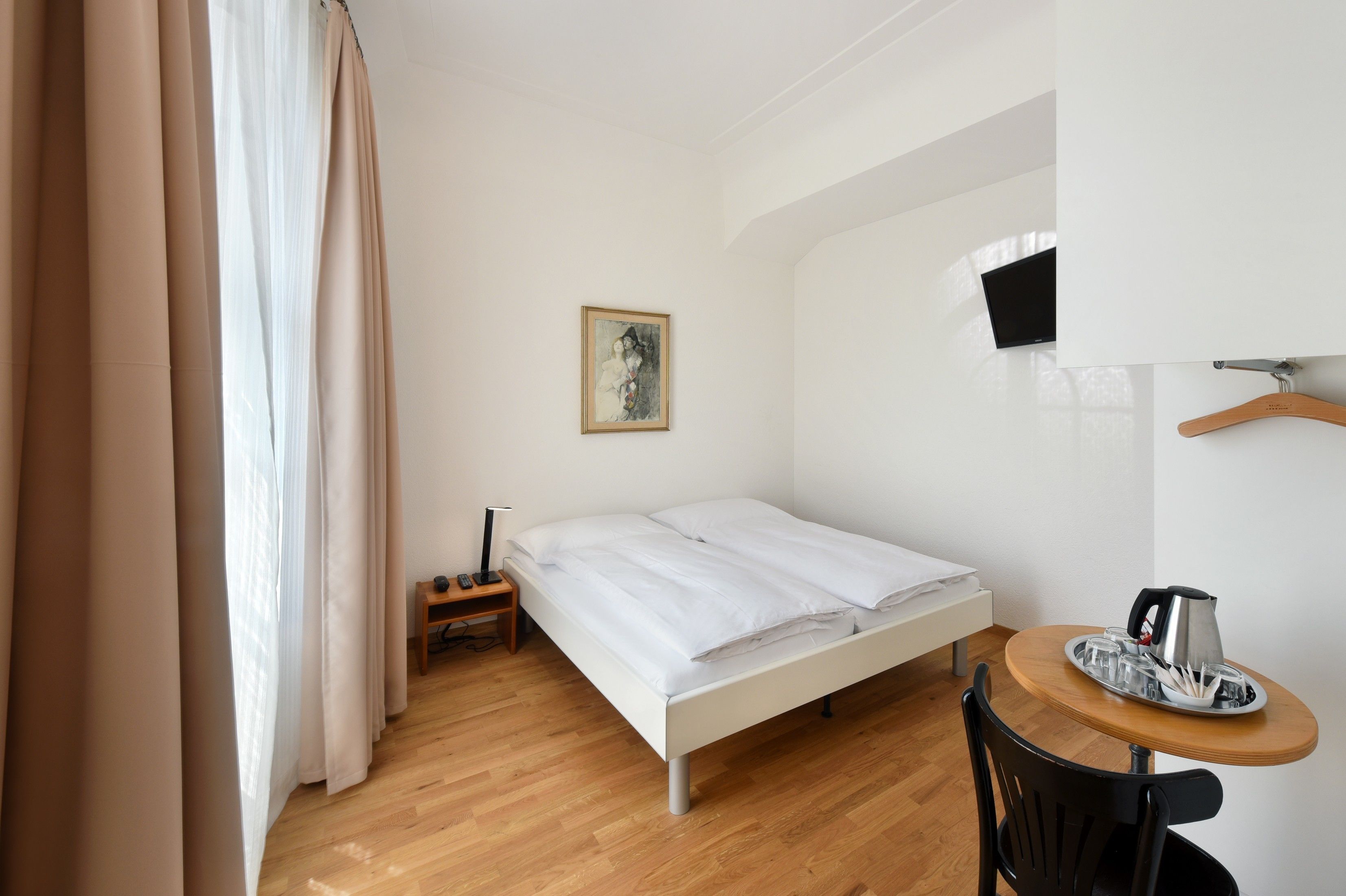 Ein kleiner, ordentlicher Raum mit einem Einzelbett, einem Nachttisch mit Lampe, einem kleinen Tisch mit Wasserkocher und einem an der Wand montierten Fernseher.