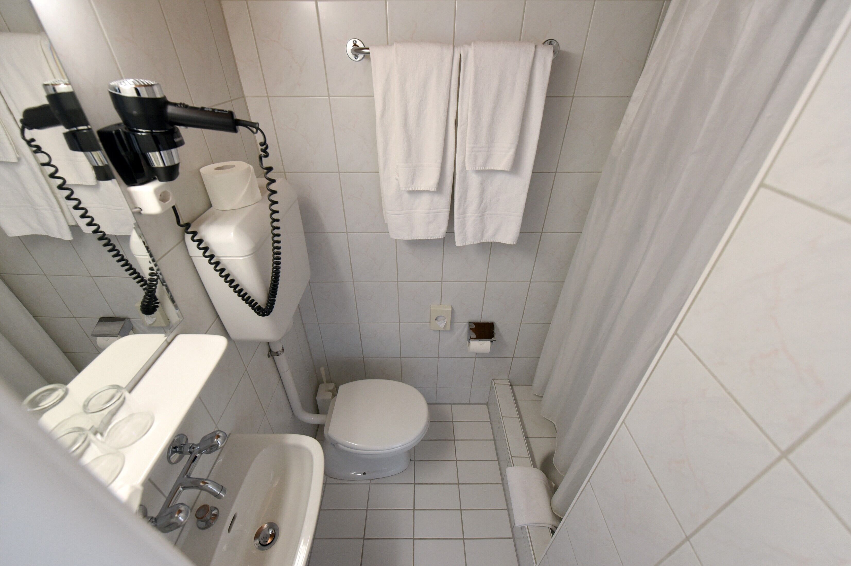A small, modern bathroom with a toilet, sink, and shower. There are towels hanging on the wall and a hairdryer on the wall.