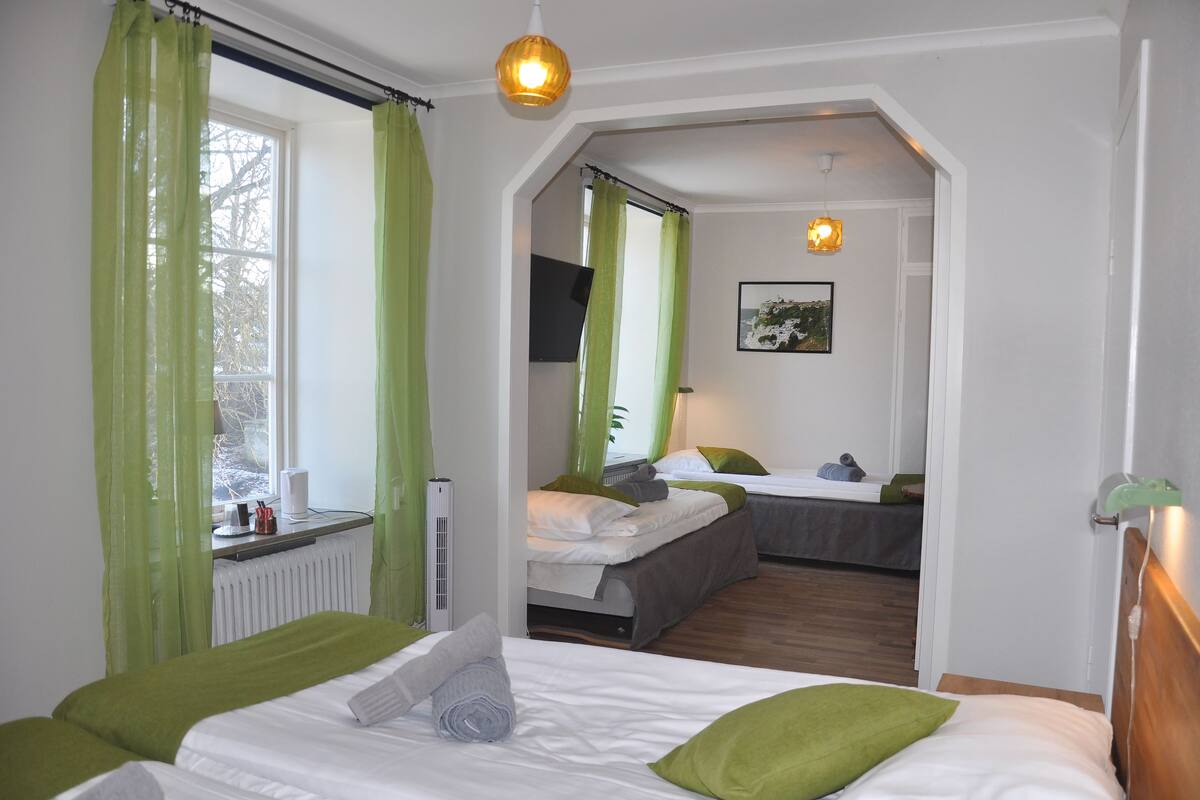 Ein Zimmer mit einem Bett, einem Fenster mit grünen Vorhängen, einem Heizkörper und einem kleinen Tisch mit einer Lampe.