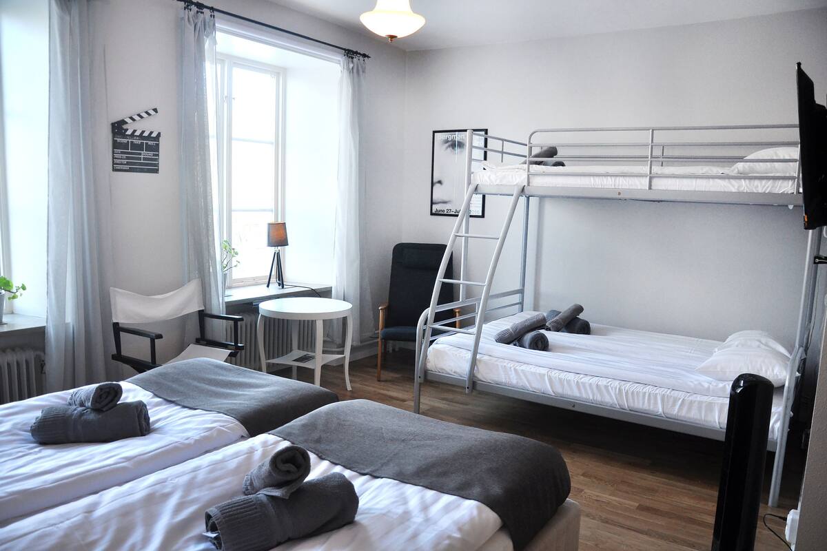 Ein Hotelzimmer mit zwei Betten, einem Etagenbett, einem kleinen Tisch, einem Stuhl und einem Fenster mit Vorhängen.