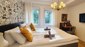 Habitación de hotel con una cama grande, dos ventanas, una araña y una televisión, además de un cuadro en la pared.