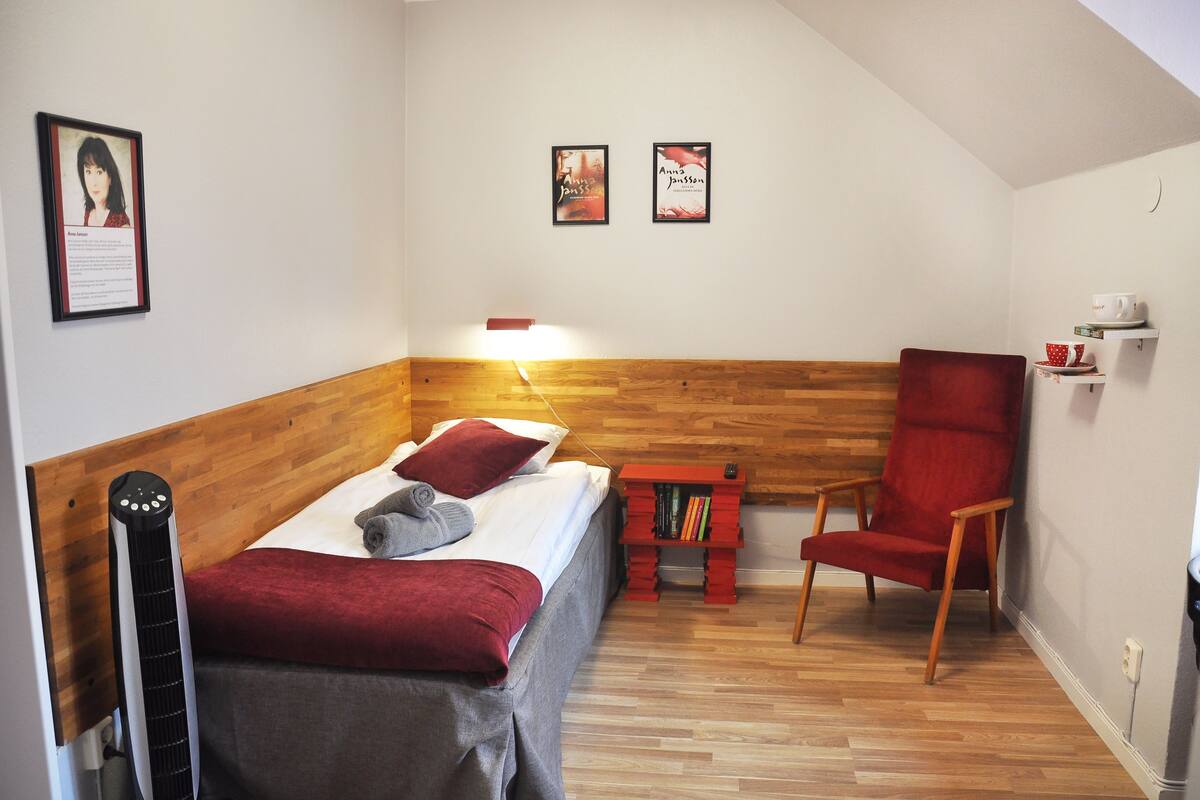 Ein kleiner, gemütlicher Raum mit einem Bett, einem roten Sessel, einem kleinen Tisch mit Büchern und zwei gerahmten Bildern an der Wand.