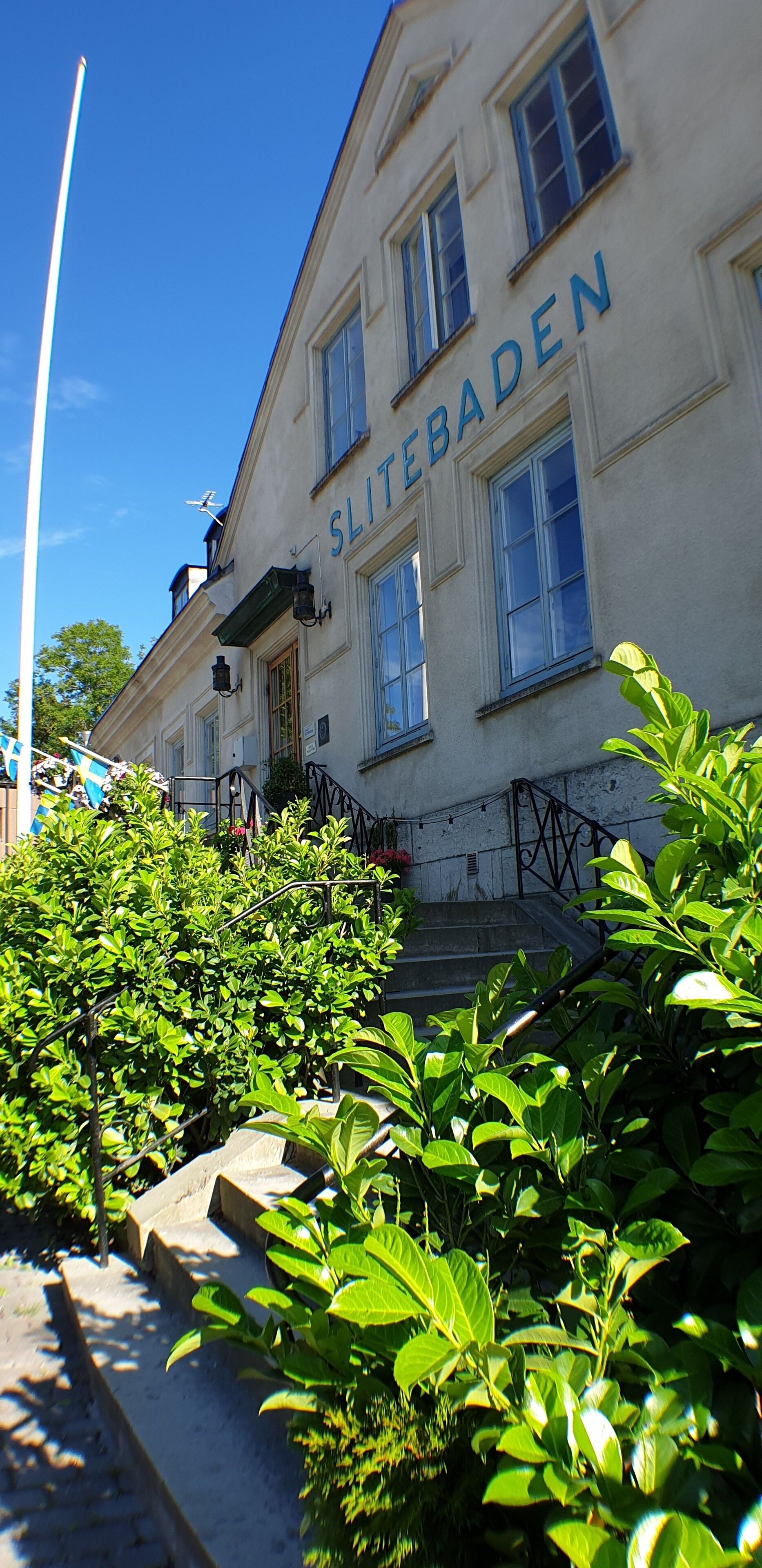 A building with the sign 'SLITEBADEN' on the facade, surrounded by lush greenery and a clear blue sky.
