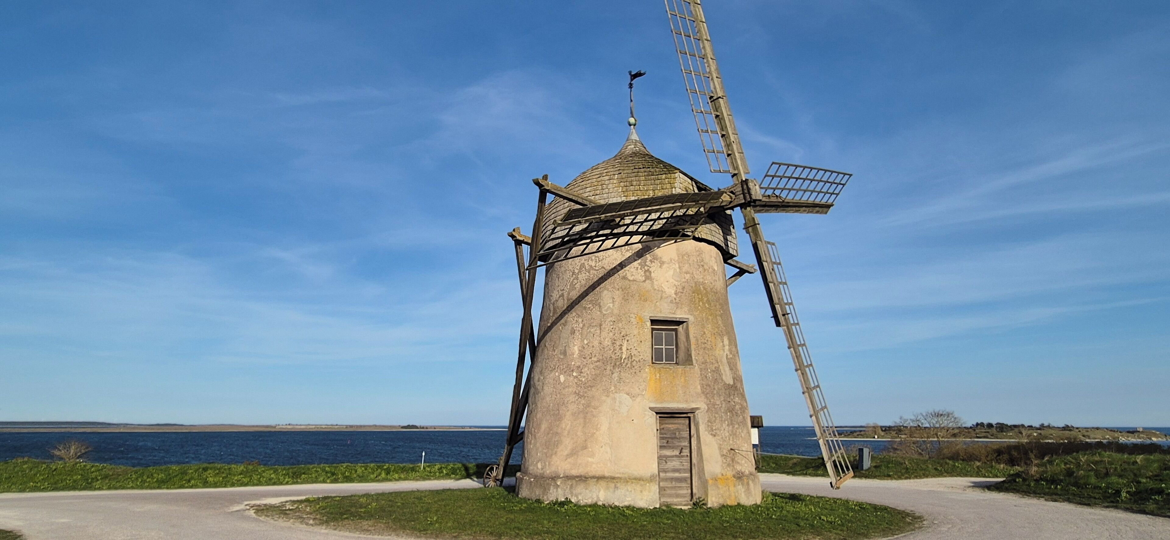 Un moulin à vent doté d’une échelle, d’une porte et d’une petite fenêtre, situé au bord d’une route d’où l’on a une vue dégagée sur la mer.