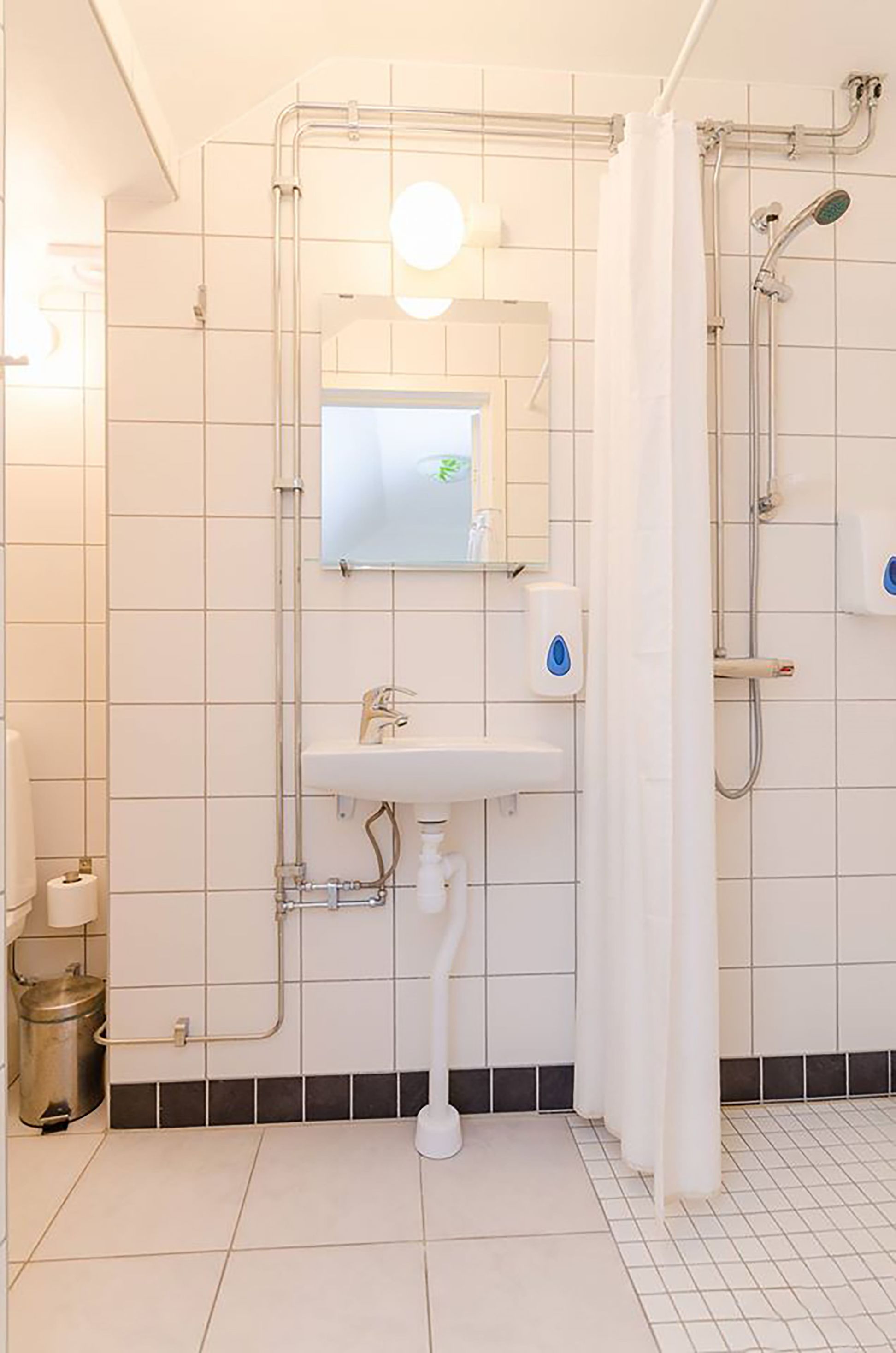 Et badeværelse med en hvid vask, spejl og brusebad. Der er en håndklædestang og en sæbedispenser.