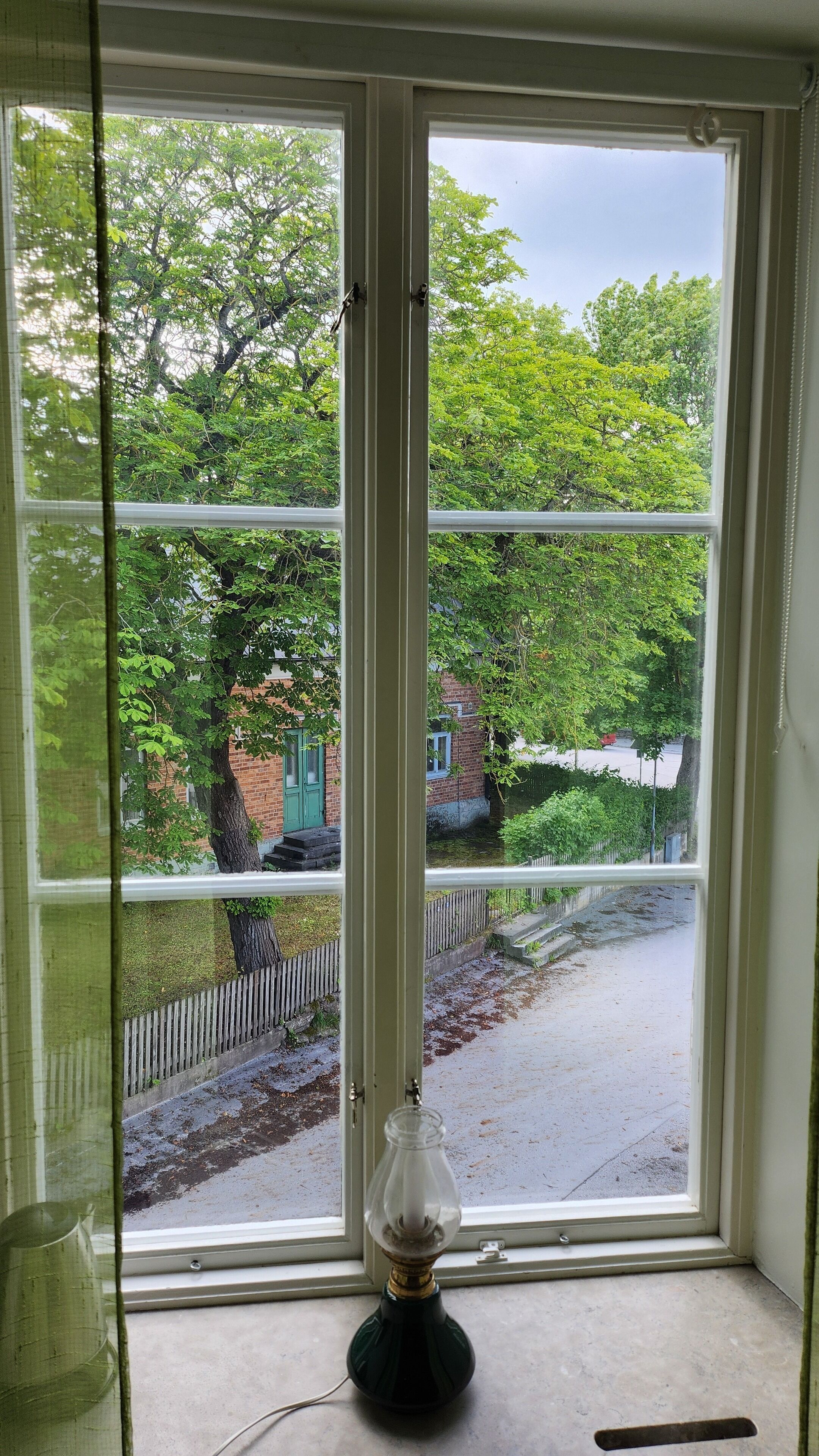 Una ventana con vista a un edificio de ladrillo y un árbol, una lámpara en el alféizar y un suelo de hormigón.