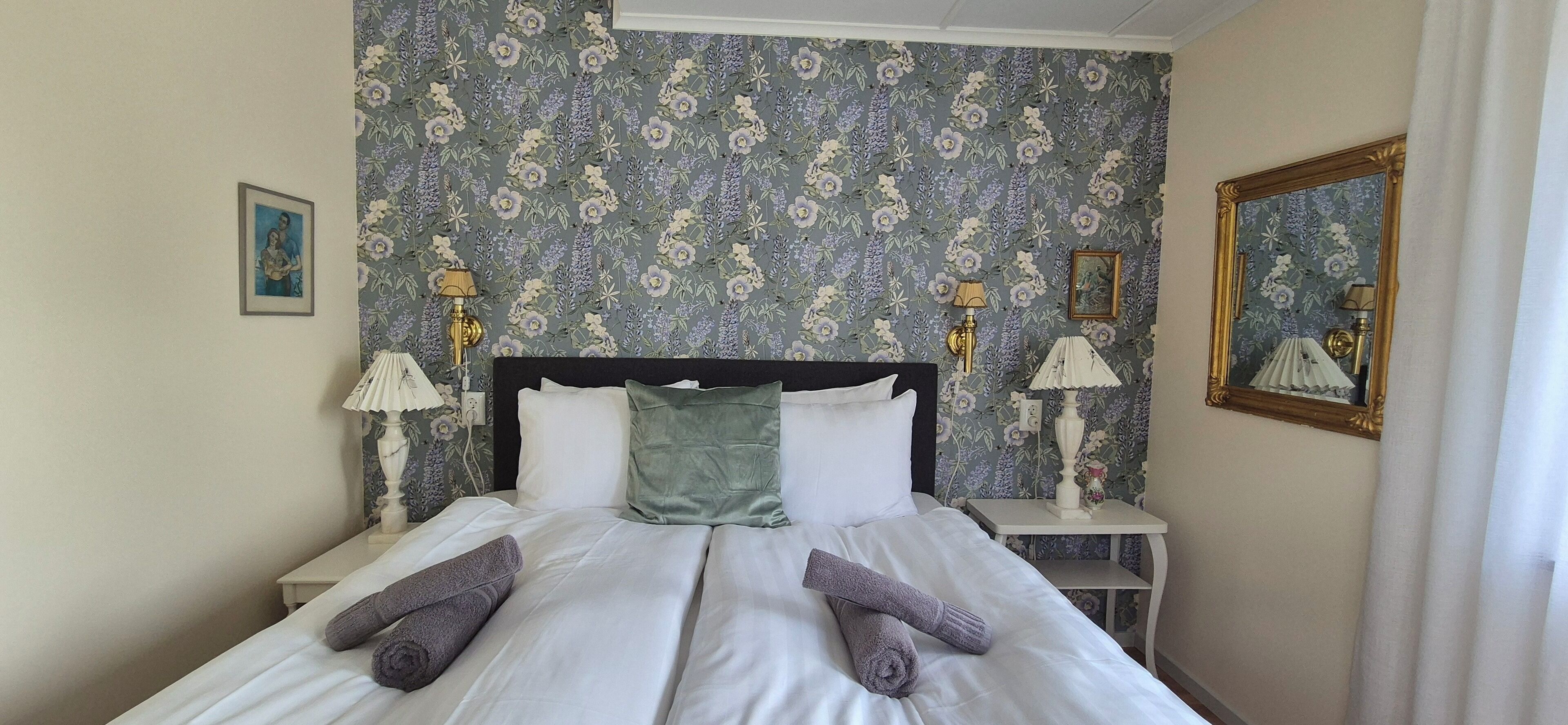 Un lit bien fait, avec du linge de lit blanc et deux serviettes enroulées, un papier peint à motifs floraux, deux lampes de chevet et un miroir encadré accroché au mur.