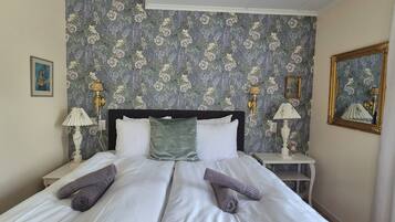 Un lit bien fait, avec du linge de lit blanc et deux serviettes enroulées, un papier peint à motifs floraux, deux lampes de chevet et un miroir encadré accroché au mur.
