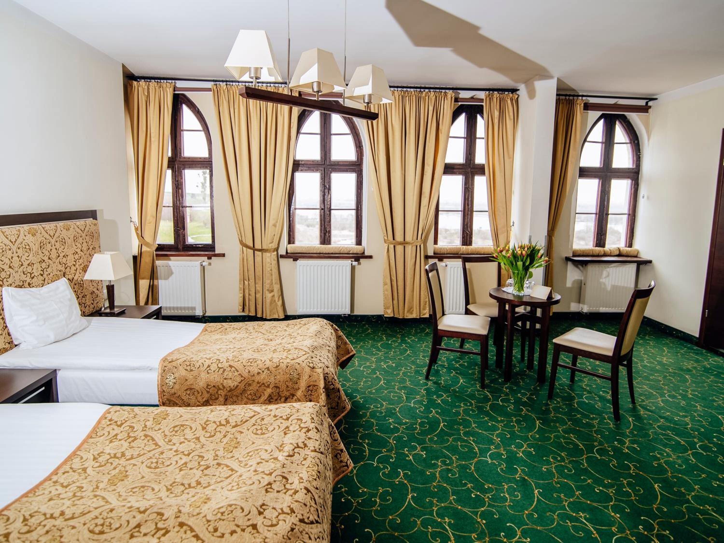 Habitación de hotel con dos camas, una mesa de comedor con sillas y ventanas grandes con cortinas.