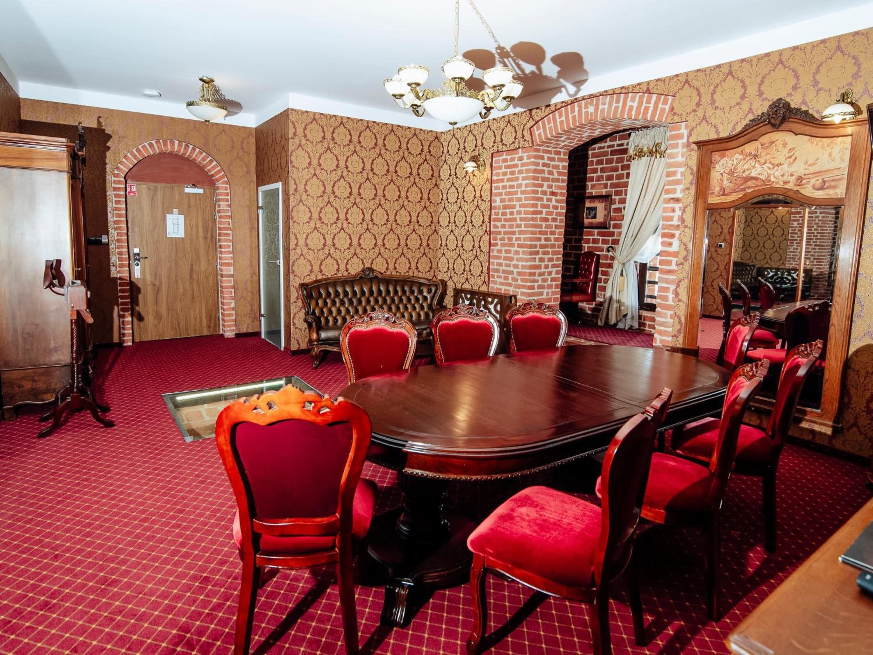 A conference room with a large round table, red upholstered chairs, a patterned wallpaper, and a brick wall.