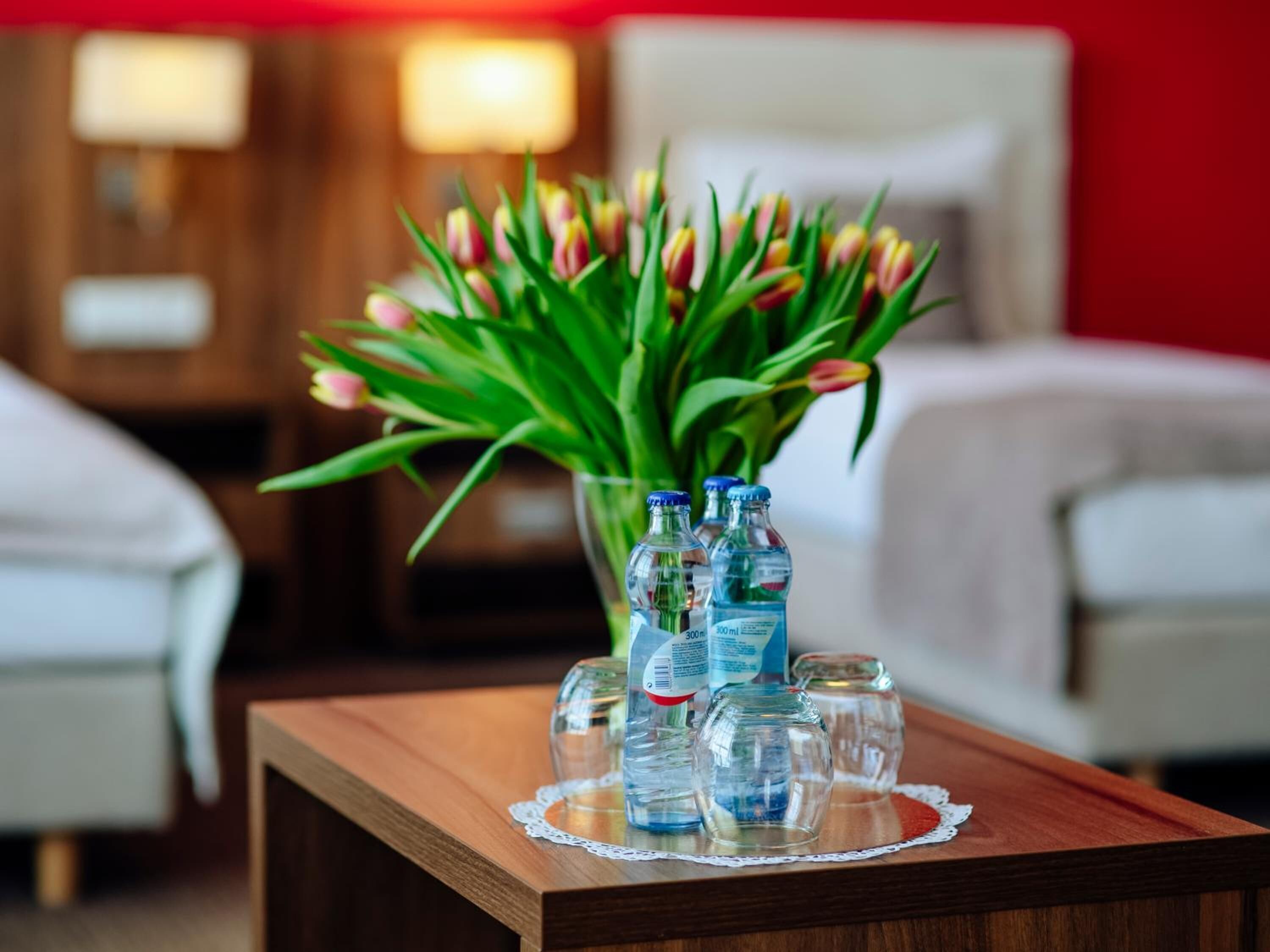 A hotel room with a wooden side table, a vase of tulips, and three water bottles on a lace doily.