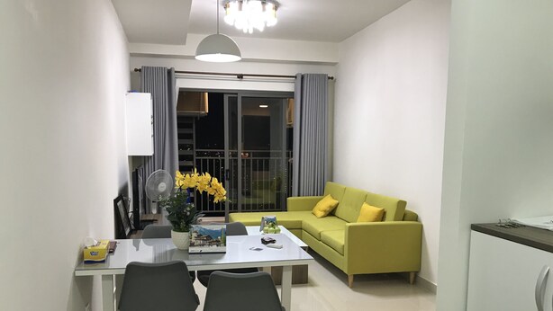 Ein modernes Wohnzimmer mit einem grünen Sofa, einem Esstisch mit zwei Stühlen, einem kleinen Tisch mit Blumen und einem großen Fenster mit Vorhängen.