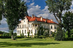 Um edifício grande e histórico com telhado de telhas vermelhas, rodeado por vegetação exuberante e árvores.