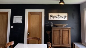 Een eetgedeelte met een witte tafel, houten stoelen, een houten kast en een bordje met de tekst 'farmhouse' aan de muur.