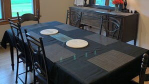 Een eetruimte met een lange tafel, stoelen, een kroonluchter, een spiegel en een raam met jaloezieën.